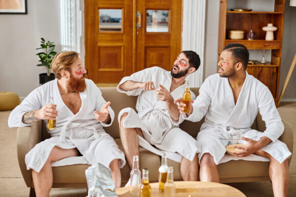 Three diverse men in bathrobes sit on the sofa, laughing and enjoying each others company in a joyful moment.