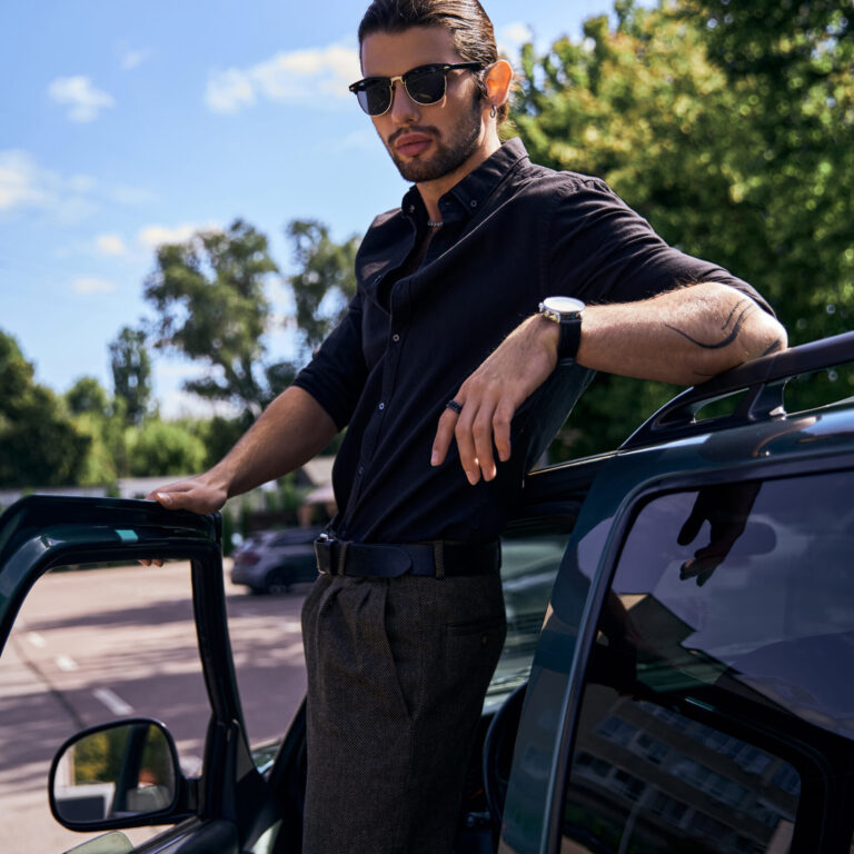 eye catching young man in stylish black outfit standing near his car and looking at camera, fashion