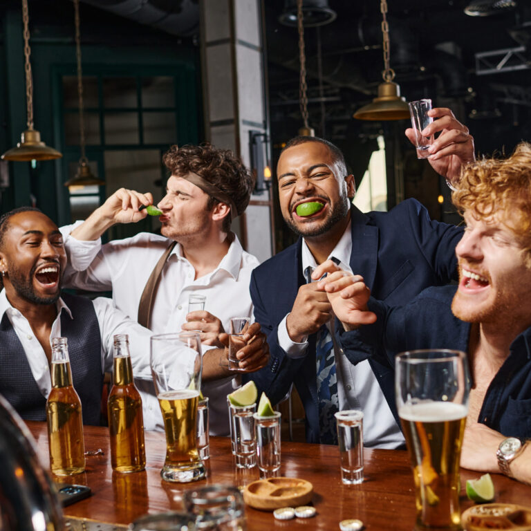 group of four happy and drunk multiethnic friends in formal wear drinking tequila in bar after work
