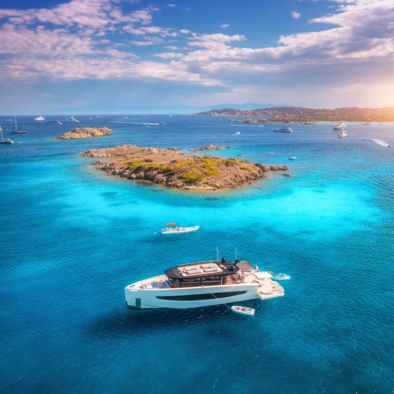 Luxury yacht on blue sea and small island at sunset in summer. Sardinia, Italy. Aerial view of boat, yachts, sea bay, transparent water and sky with clouds. Top view of seaside. Tropical seascape